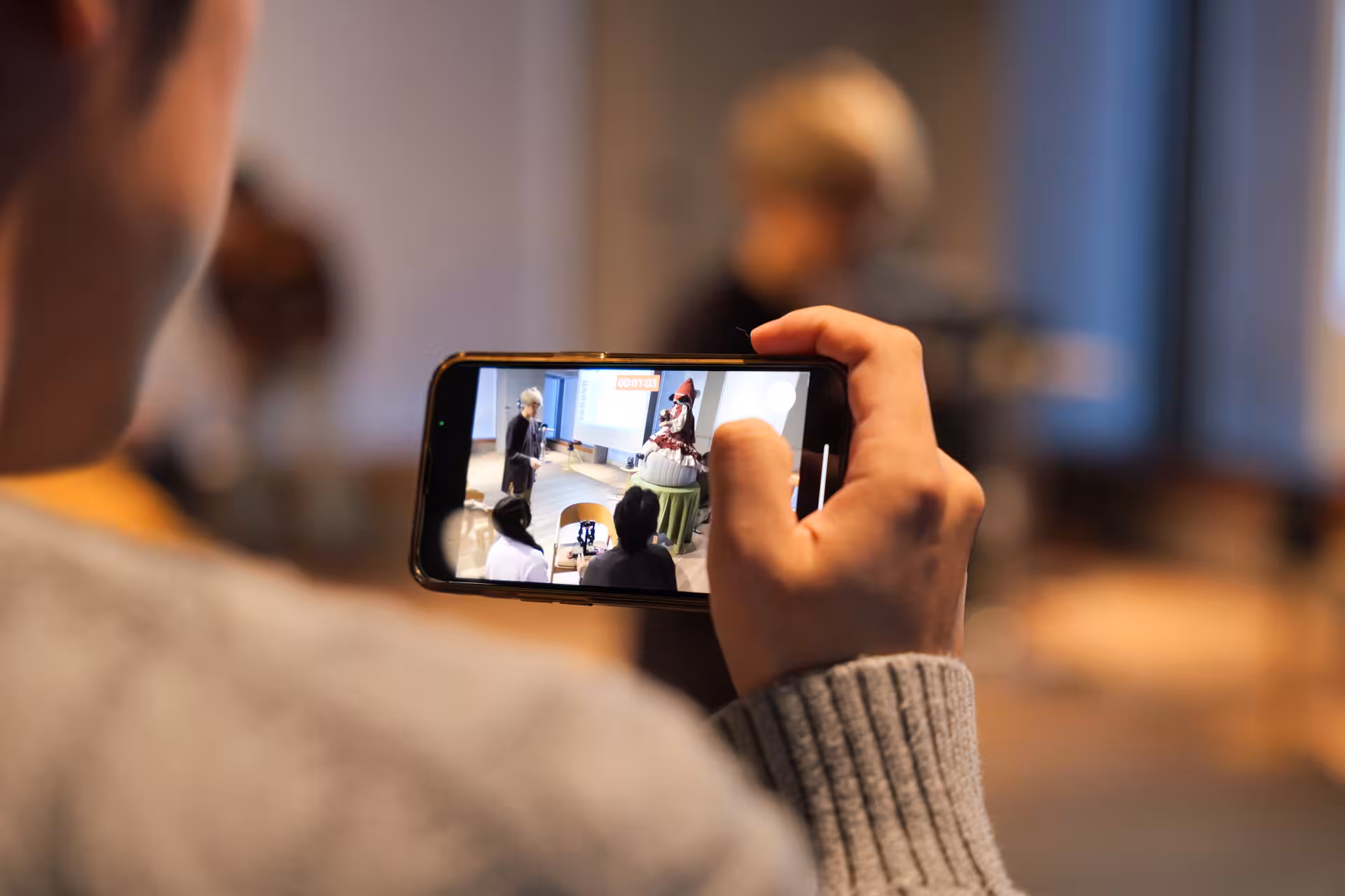 Close-up of someone's smartphone camera filming as people are next to a doll seated on a round cushion on a table