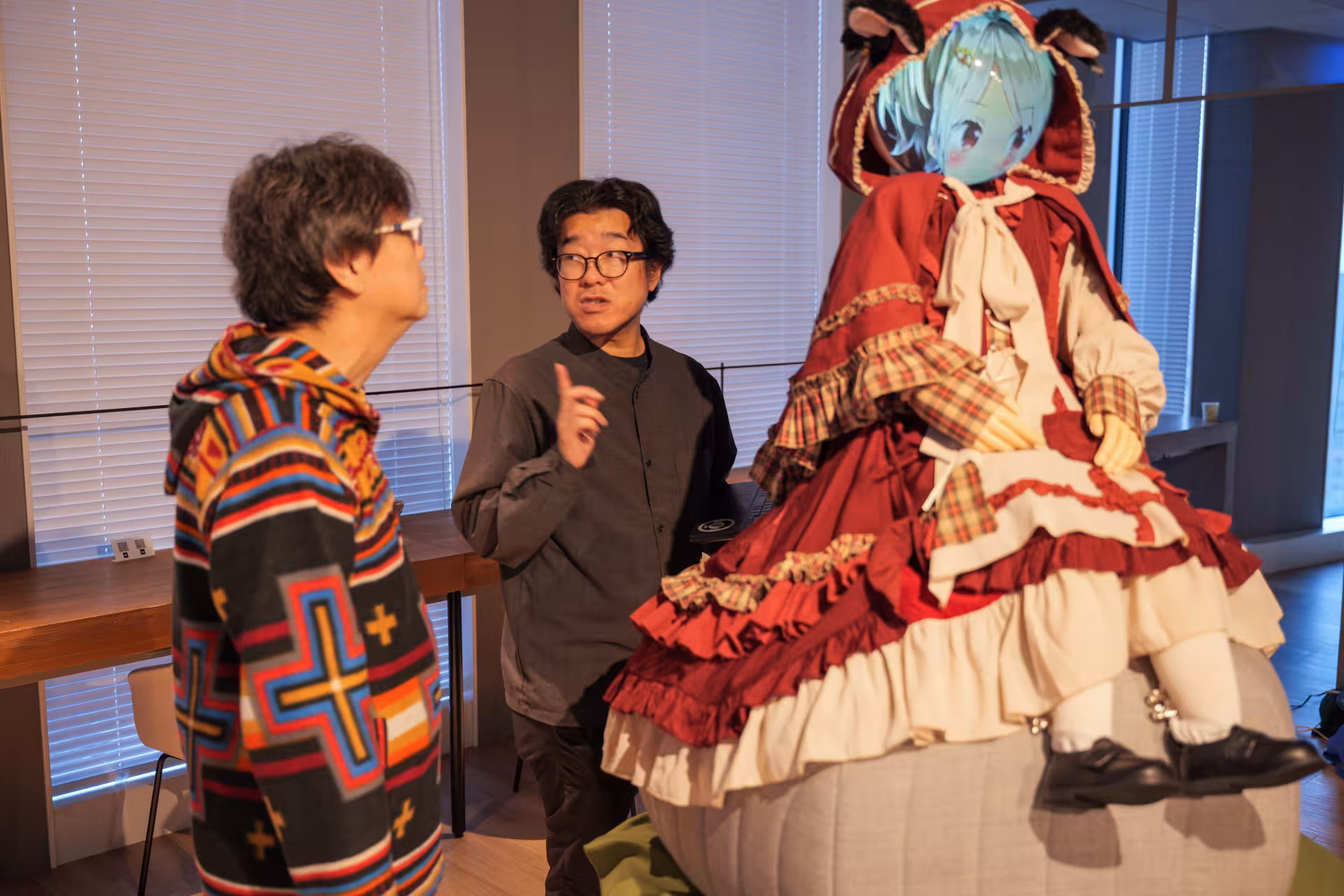 Two men in conversation next to a tall doll seated on a big round cushion on a table