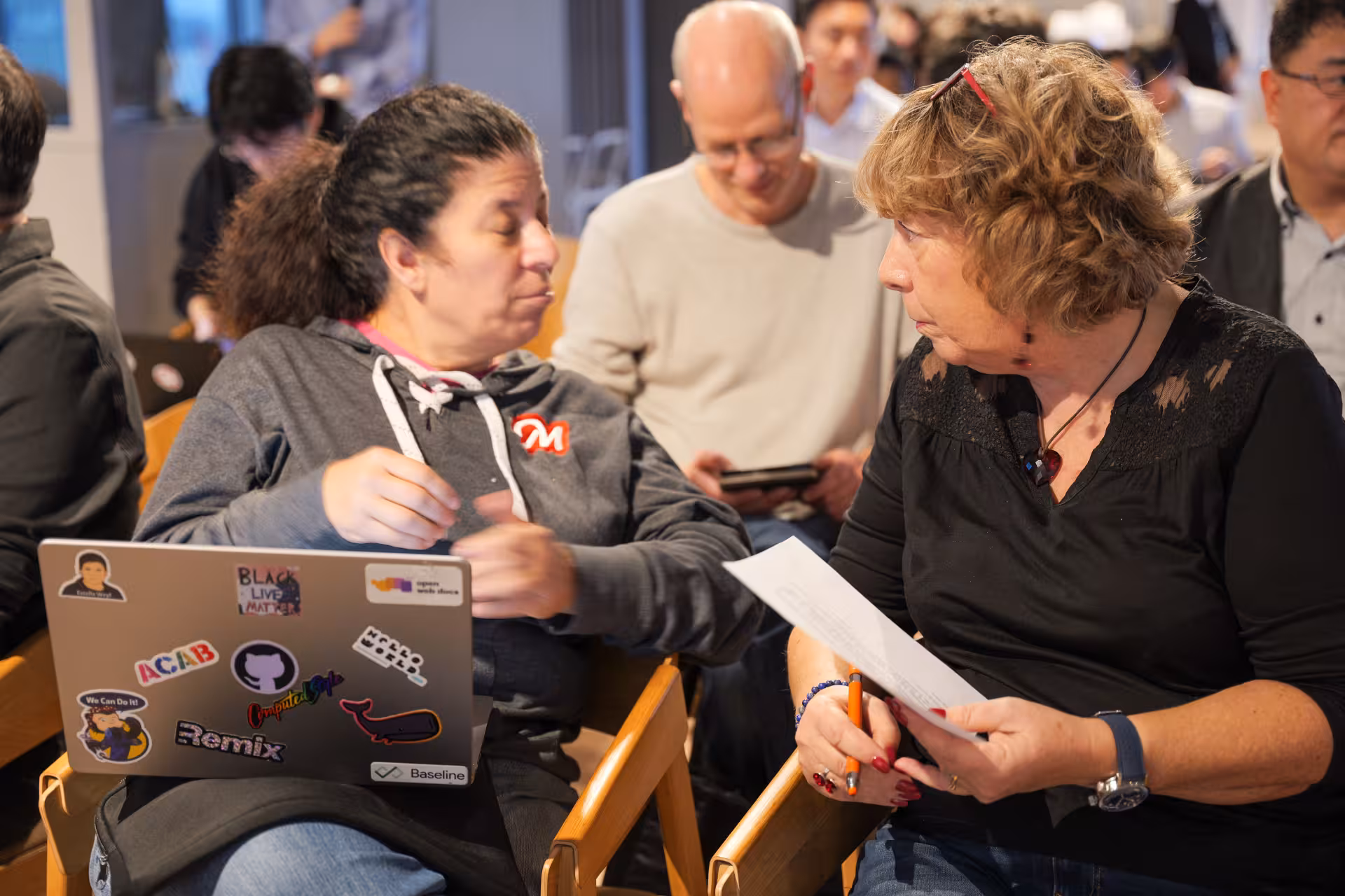 Two women on chairs talking to each other. One of them has an open computer on her lap, the other holds a sheet of paper and a pen. More people are visible in the background