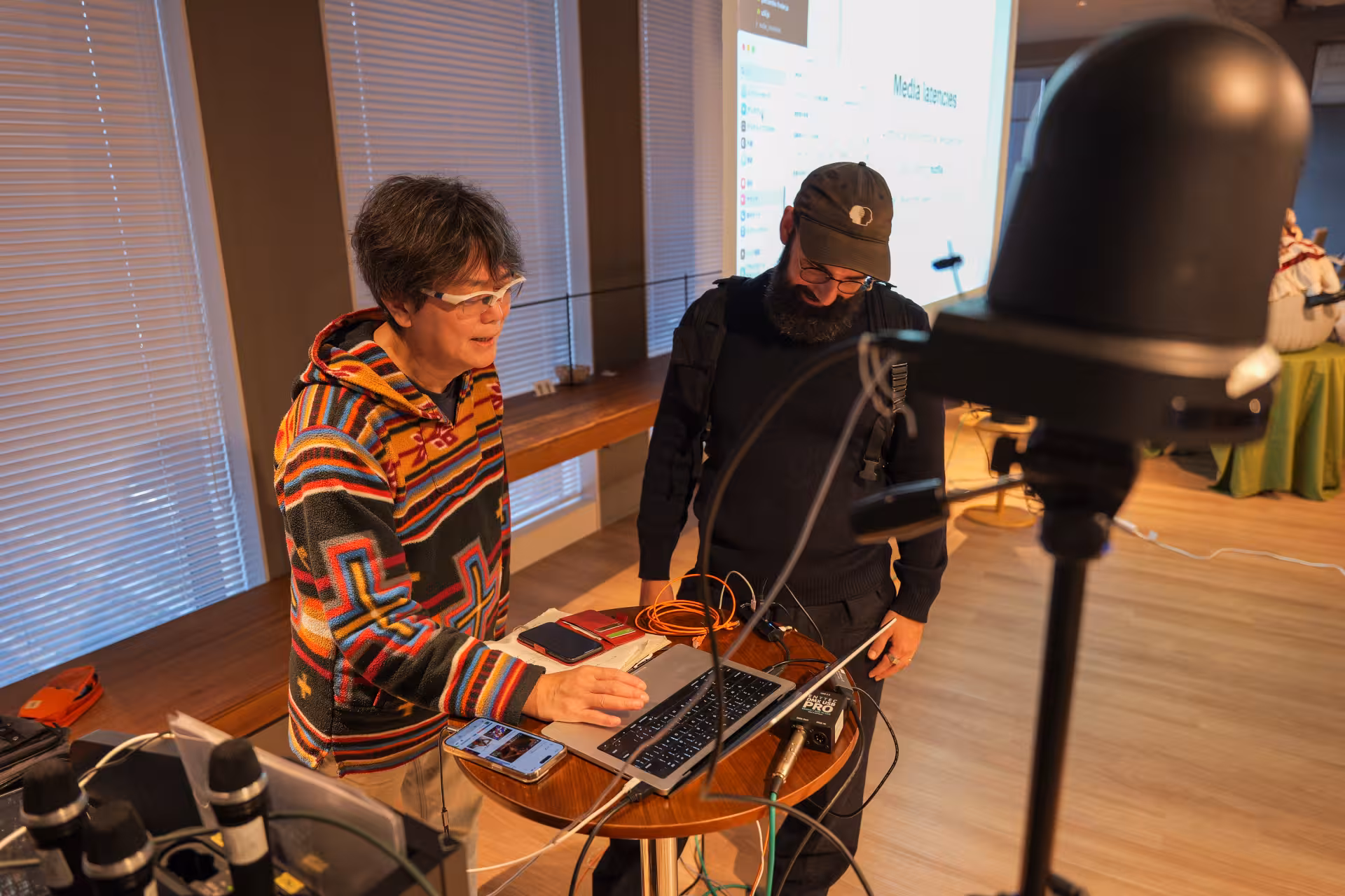 Two men. One typing at a laptop connected to many other devices, the other watching the laptop's screen
