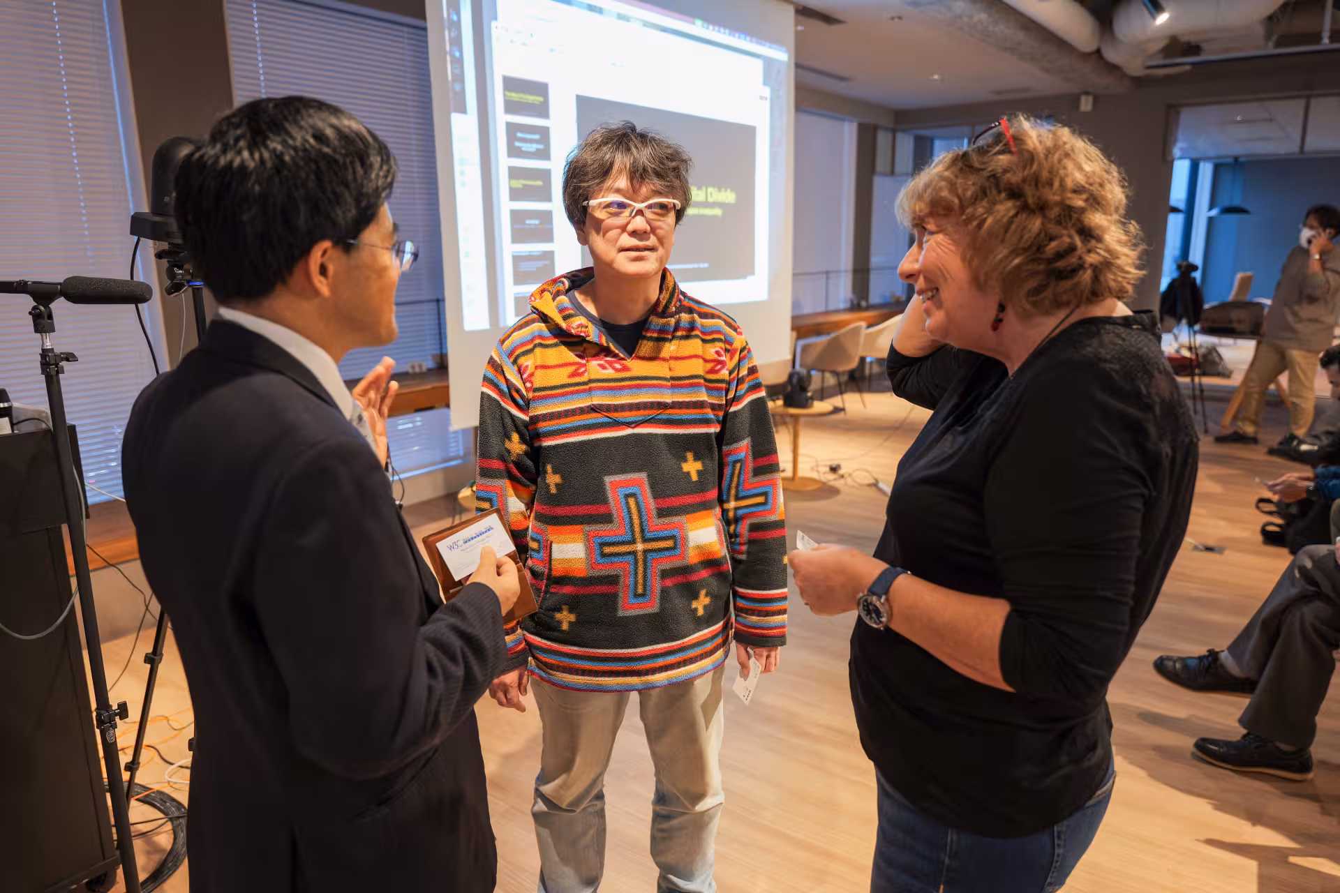 Three persons in a conversation in a room where a large white screen is deployed and various people look at it.